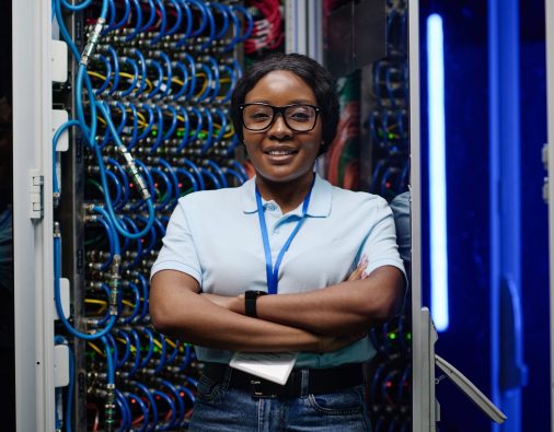 Portrait of African female engineer smiling at camera standing with her arms crossed in data server with cables in rack in background